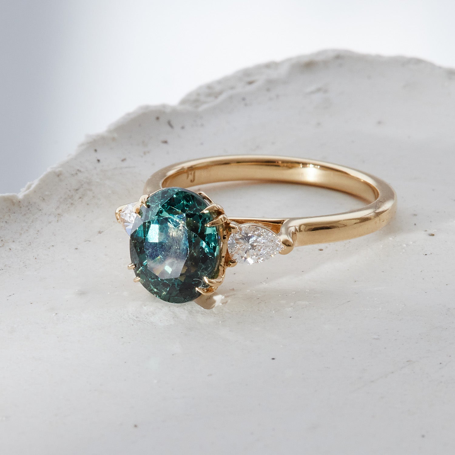 Sheree trilogy engagement ring featuring a teal, round-cut sapphire flanked by pear-cut white diamonds in yellow gold band
