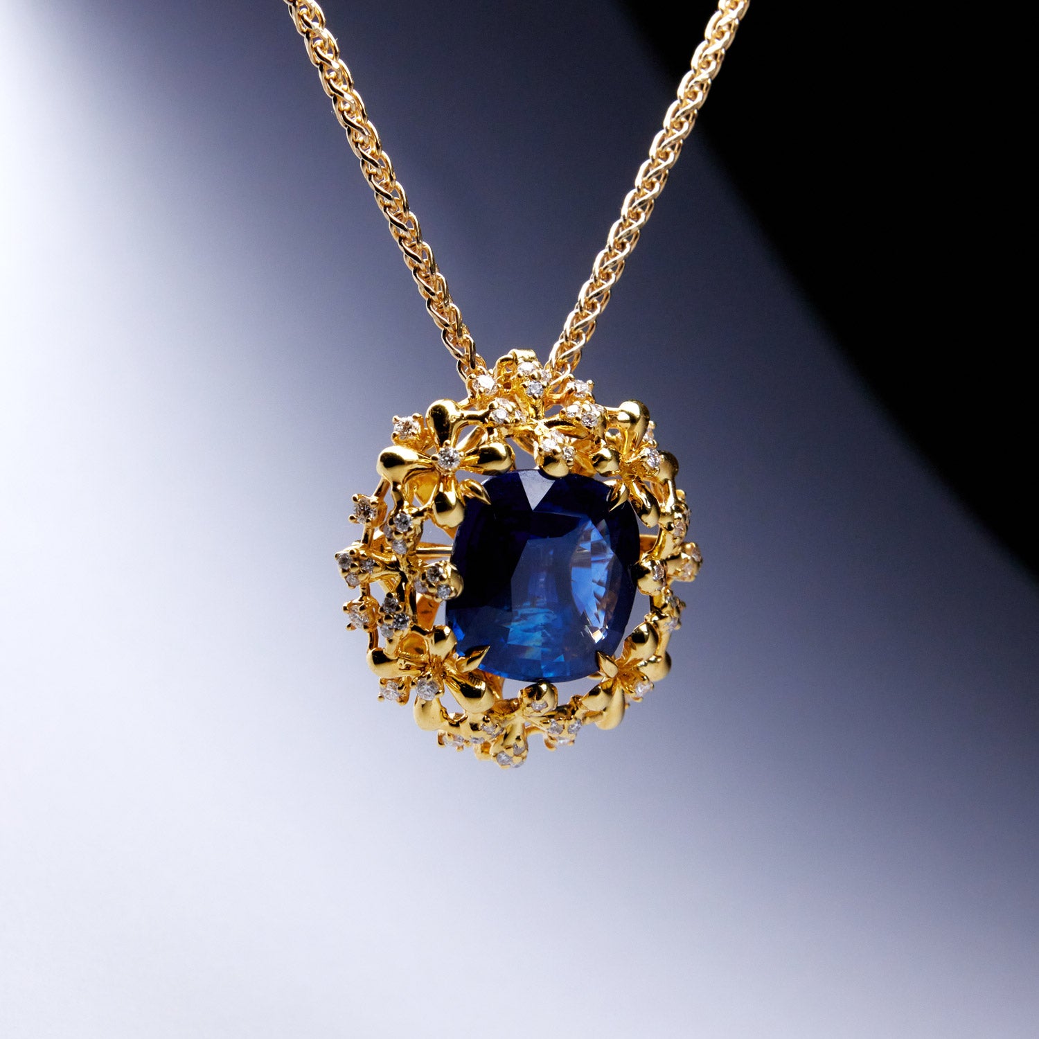 A luxurious interchangeable necklace and ring set featuring a royal blue Ceylon sapphire, surrounded by a floral wreath of golden florals with white diamonds, all in 18k yellow gold.