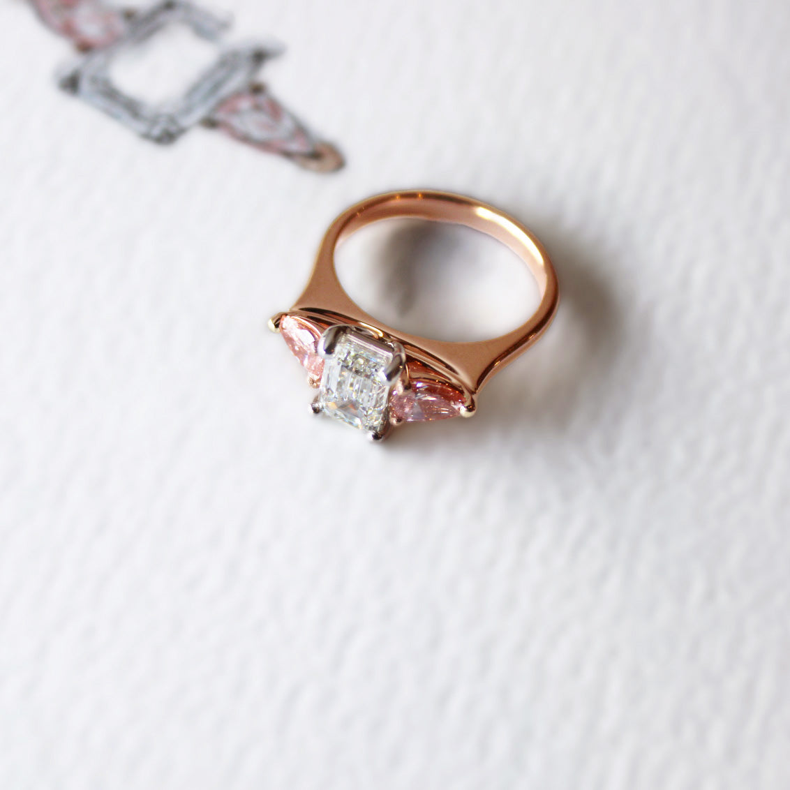 Bespoke trilogy engagement ring in 18k rose gold with emerald-cut diamond centre and pink diamond accents, elegant and timeless design.