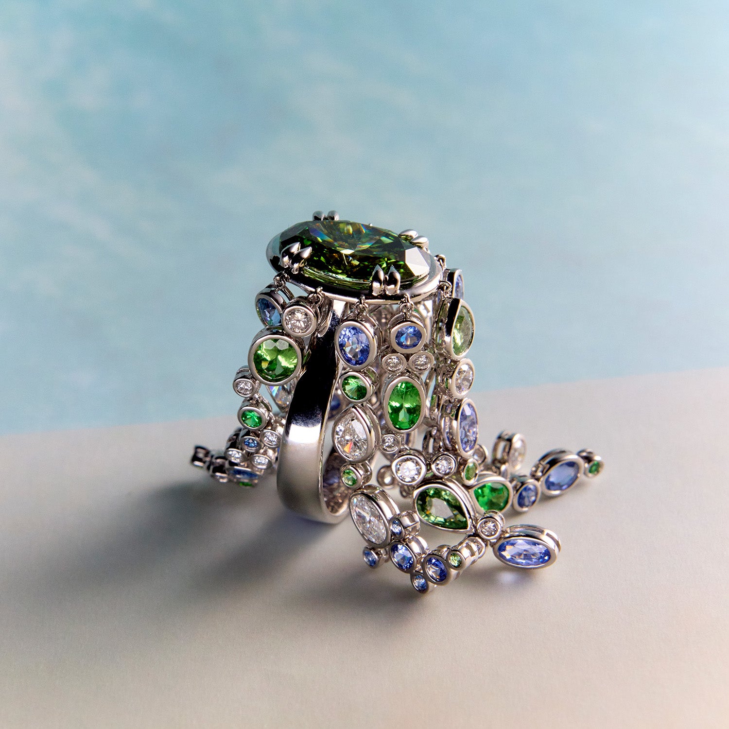 Waterfall ring with streams of blue sapphires, green sapphires, and white diamonds cascade organically across the hand, echoing the rhythm of water catching light as it moves over stone on a light blue background