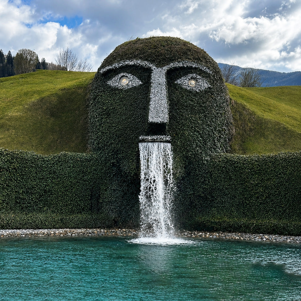 Ann Holiday waterfall with garden sculpture of a face with a waterfall coming out of the mouth, set against a scenic background with mountains and clouds.
