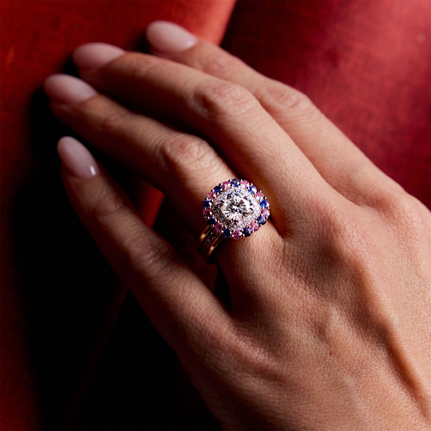 Yellow gold sapphire and diamond ring with pink and blue halo setting, worn on hand against red background