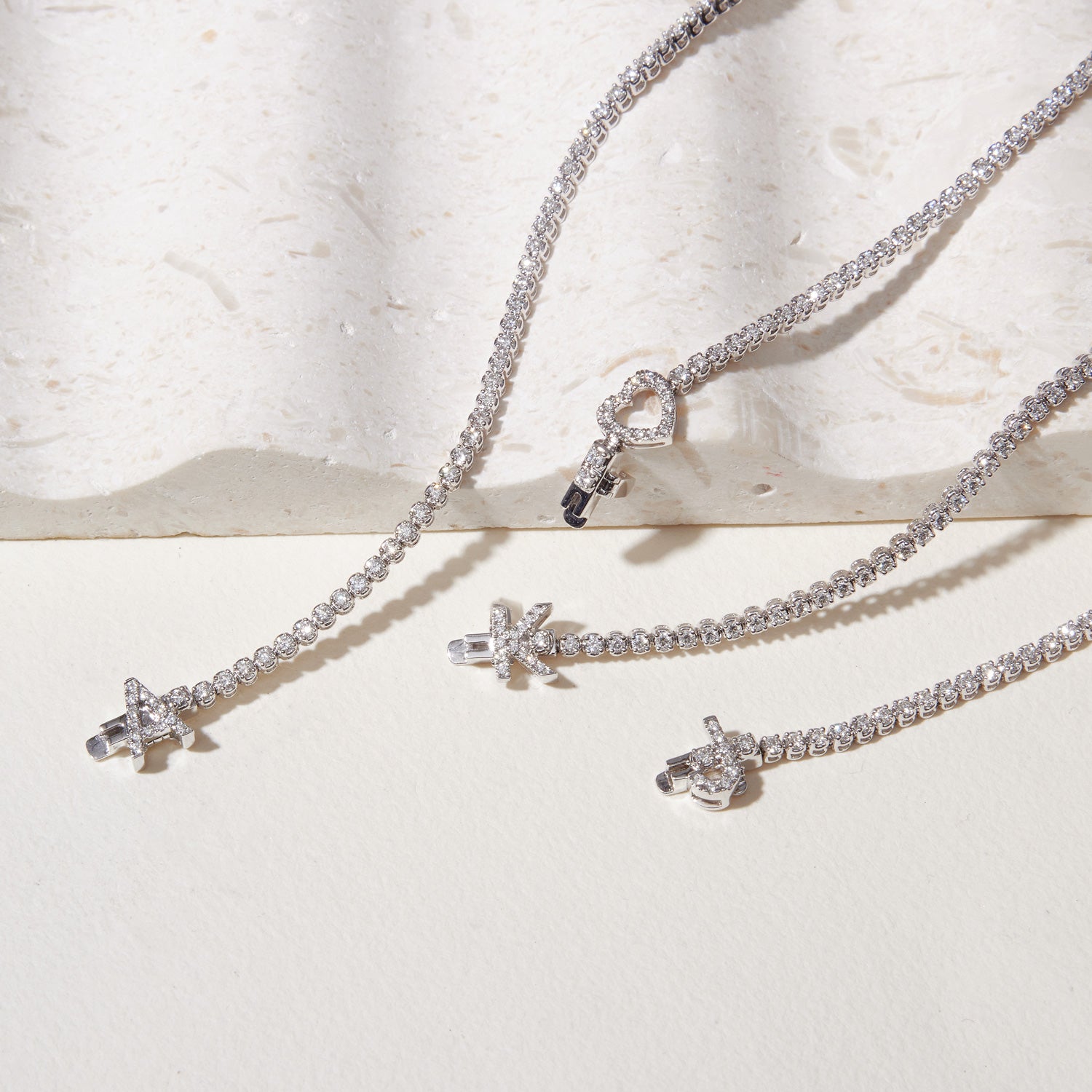 Faith Jewels bespoke initial diamond tennis bracelets in white gold, each featuring a unique clasp shaped as a letter or symbol — including X, A, K, and a heart — set with round brilliant-cut diamonds
